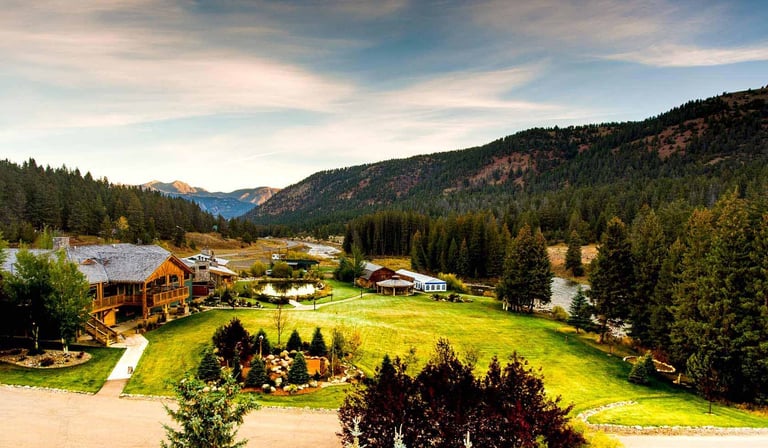 Aerial view of a rural mountain valley with wooden cabins, green meadows, evergreen forests, and a river winding through the landscape