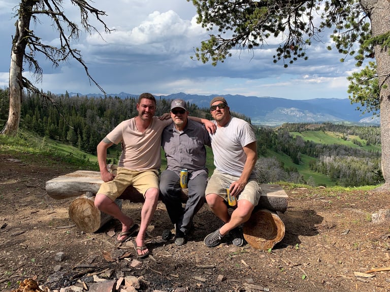 Three people sitting on a log at a mountain overlook with forested valley landscape in background