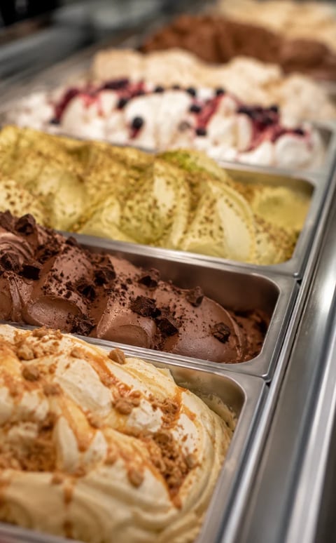 Multiple flavors of gelato in metal display pans including pistachio, chocolate, berry, and cream varieties