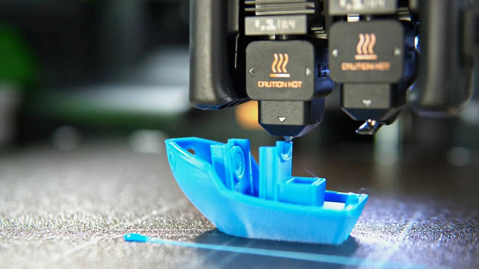 3D printer nozzles creating a small blue plastic boat on a metal build platform