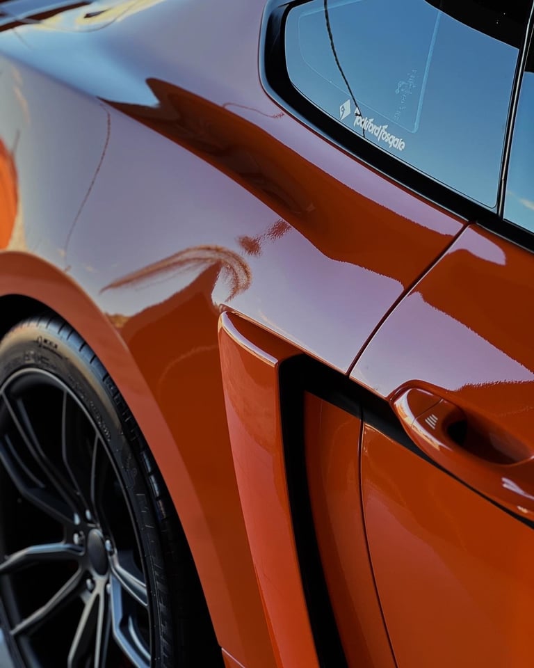 Close-up of a shiny red or orange sports car body with window and black alloy wheel visible against blue sky