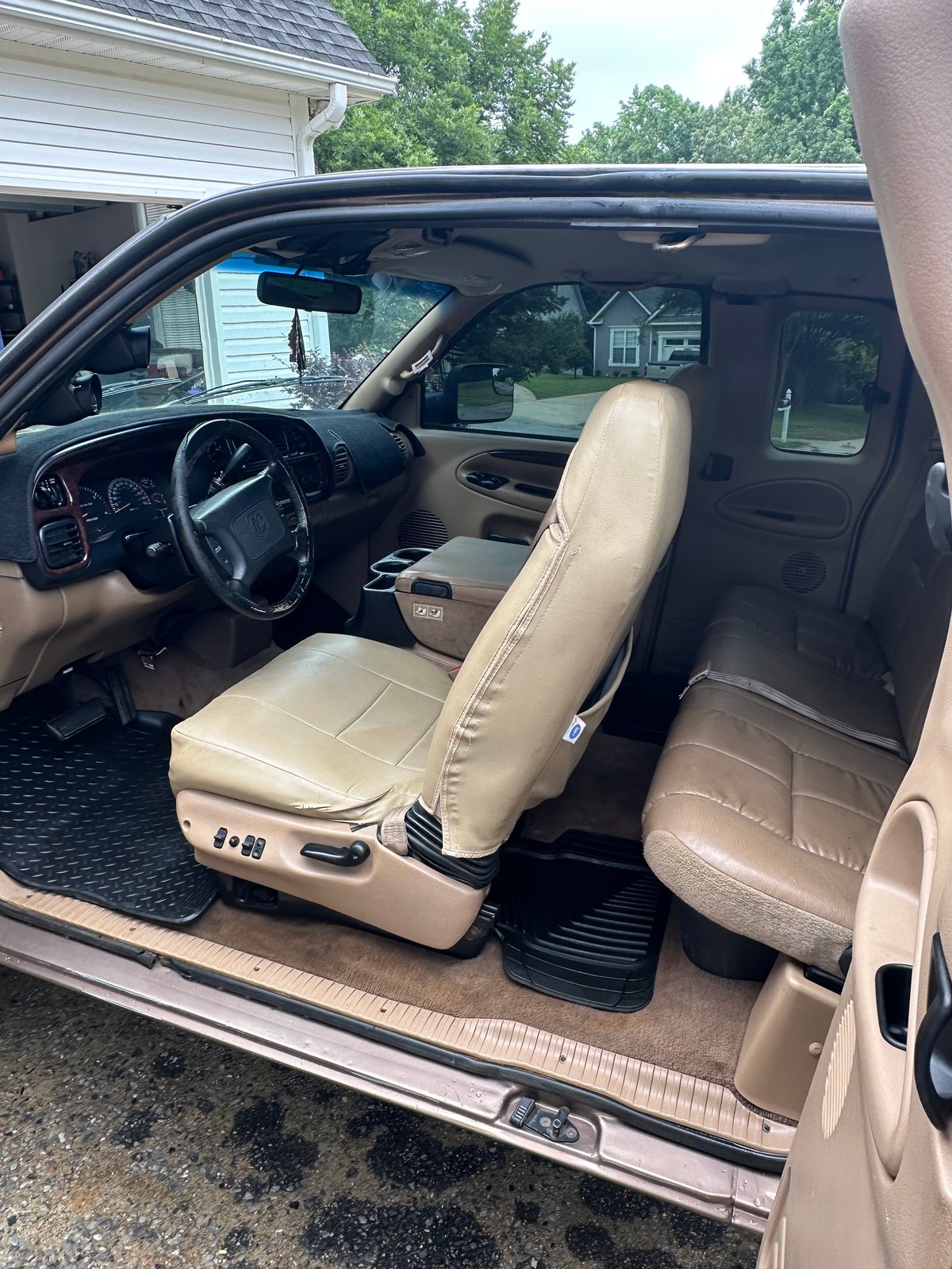 Interior view of a classic truck with tan leather seats, steering wheel, and dashboard, door open with residential driveway in background