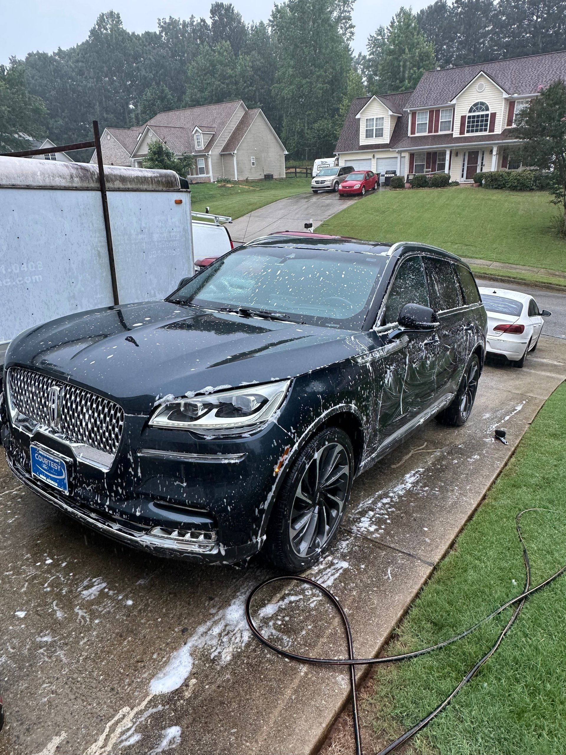 Black SUV being washed in residential driveway with suburban homes and trees in background
