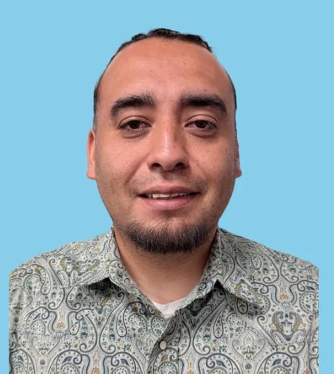Headshot of a man with dark hair and beard smiling at camera wearing a patterned button-up shirt against a blue background