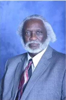 Professional headshot of Hayy Oliver wearing a gray suit jacket and red tie against a blue background