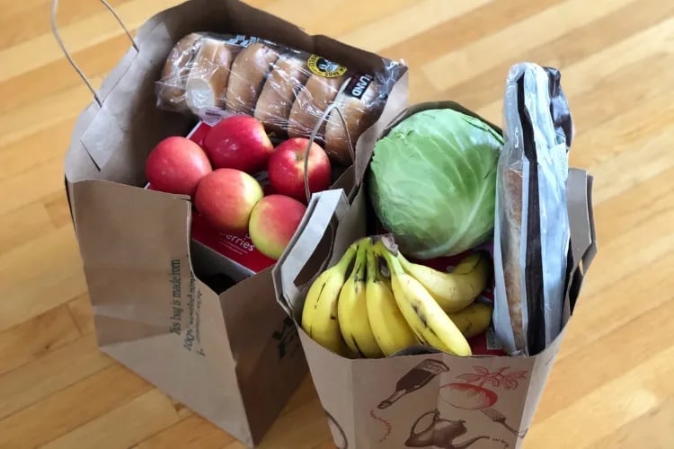 Two cardboard boxes filled with fresh groceries including red apples, bananas, green cabbage, and packaged items on a wooden floor