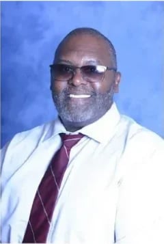 Professional headshot of Moses George wearing glasses, white coat, and purple tie against a blue background