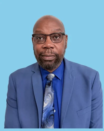 Professional headshot of a Black man wearing glasses, a blue blazer, and blue tie against a light blue background