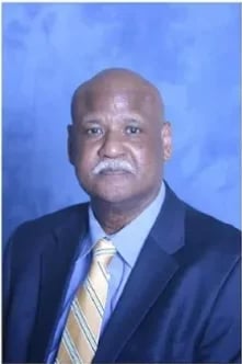 Professional headshot of Amos Carpenter wearing a dark blue suit and striped tie against a blue background