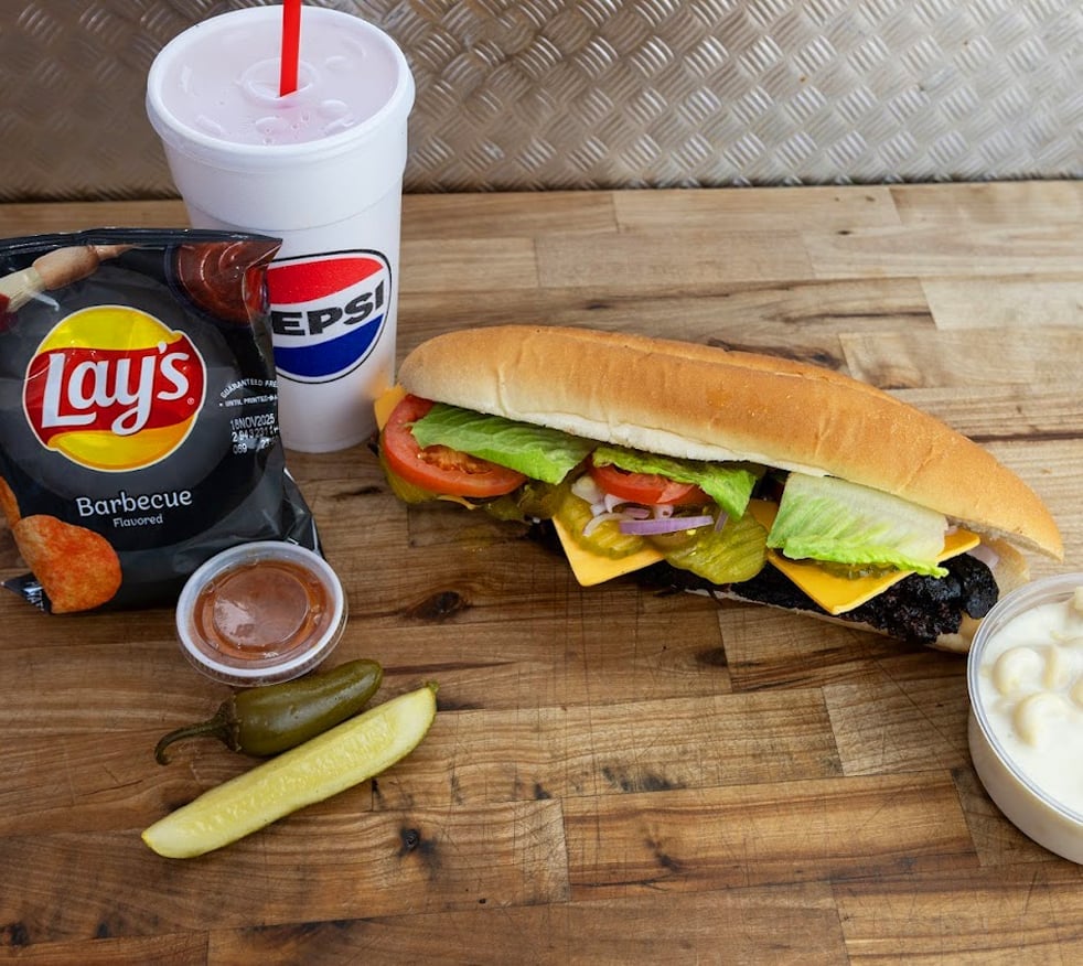 Meal spread on wooden table with submarine sandwich, Lays barbecue chips, Pepsi drink, pickle, sauce, and dip
