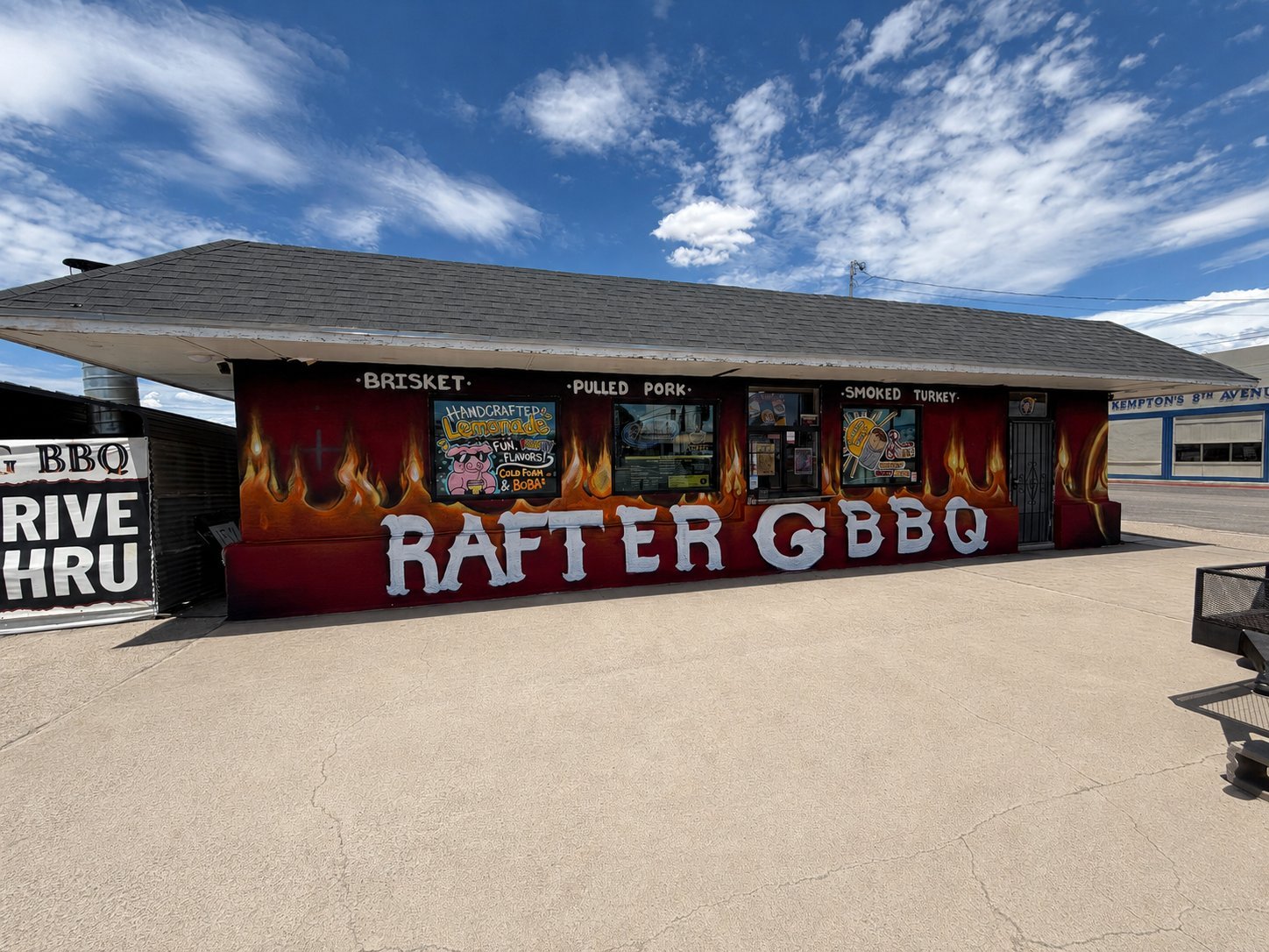 Red Rafter G BBQ building with flame graphics and signage advertising brisket, pulled pork, and smoked turkey, in a desert parking lot under a blue sky with white clouds