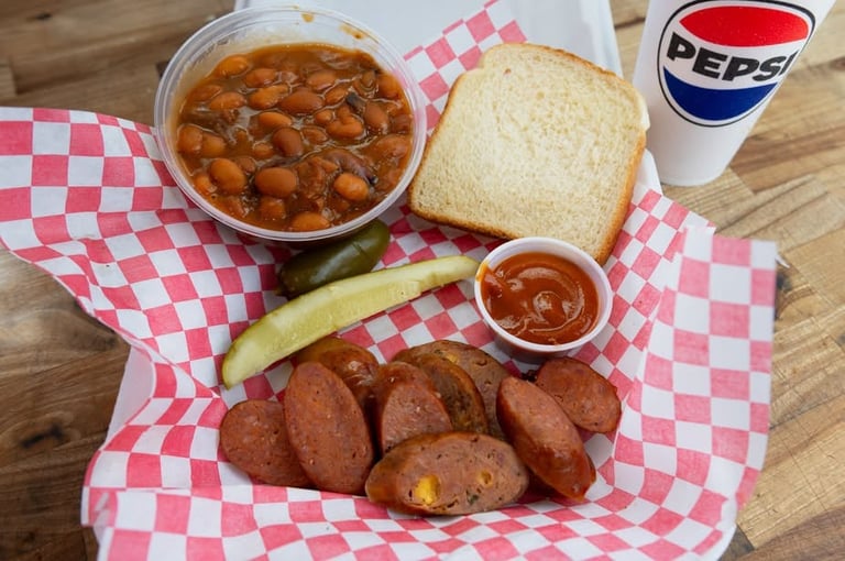 Sliced smoked sausage on a plate with baked beans, white bread, pickle, and BBQ sauce