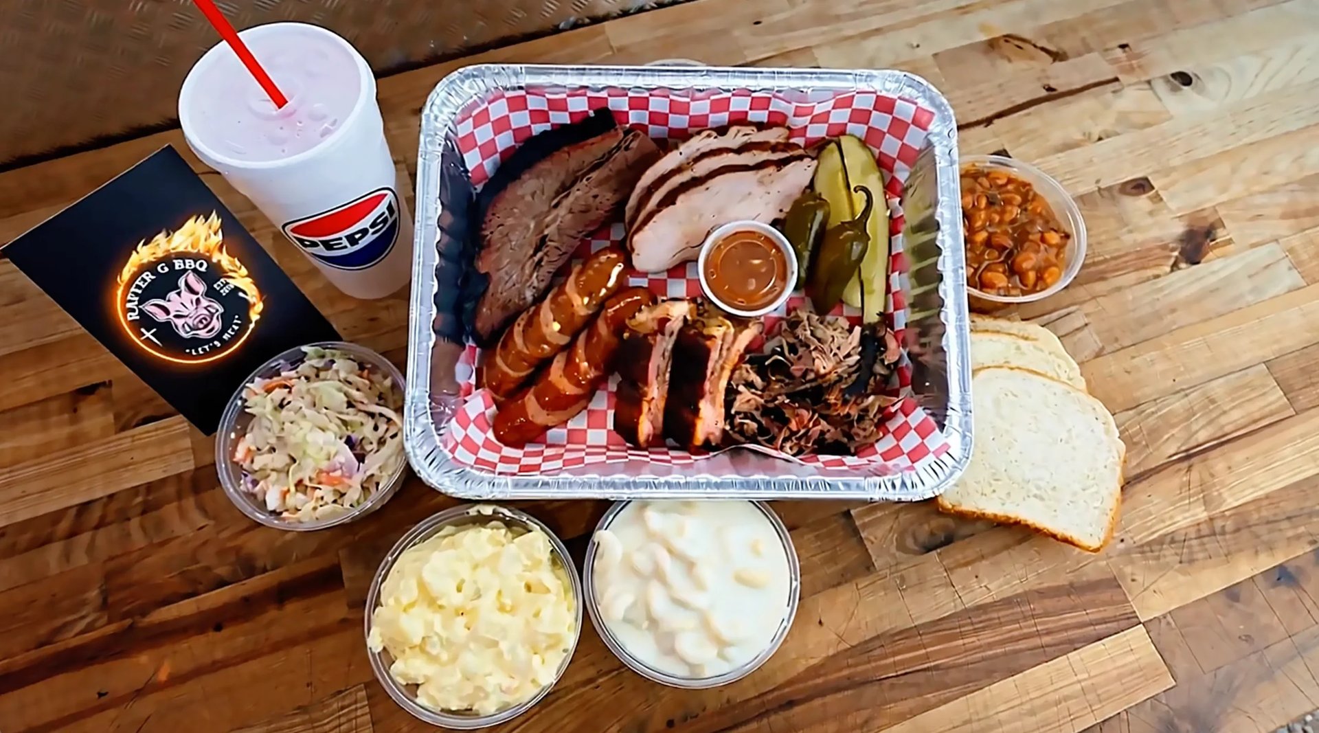 Overhead view of a Rafter G BBQ spread — meats, sauce, coleslaw, sides, and a drink on a wooden table