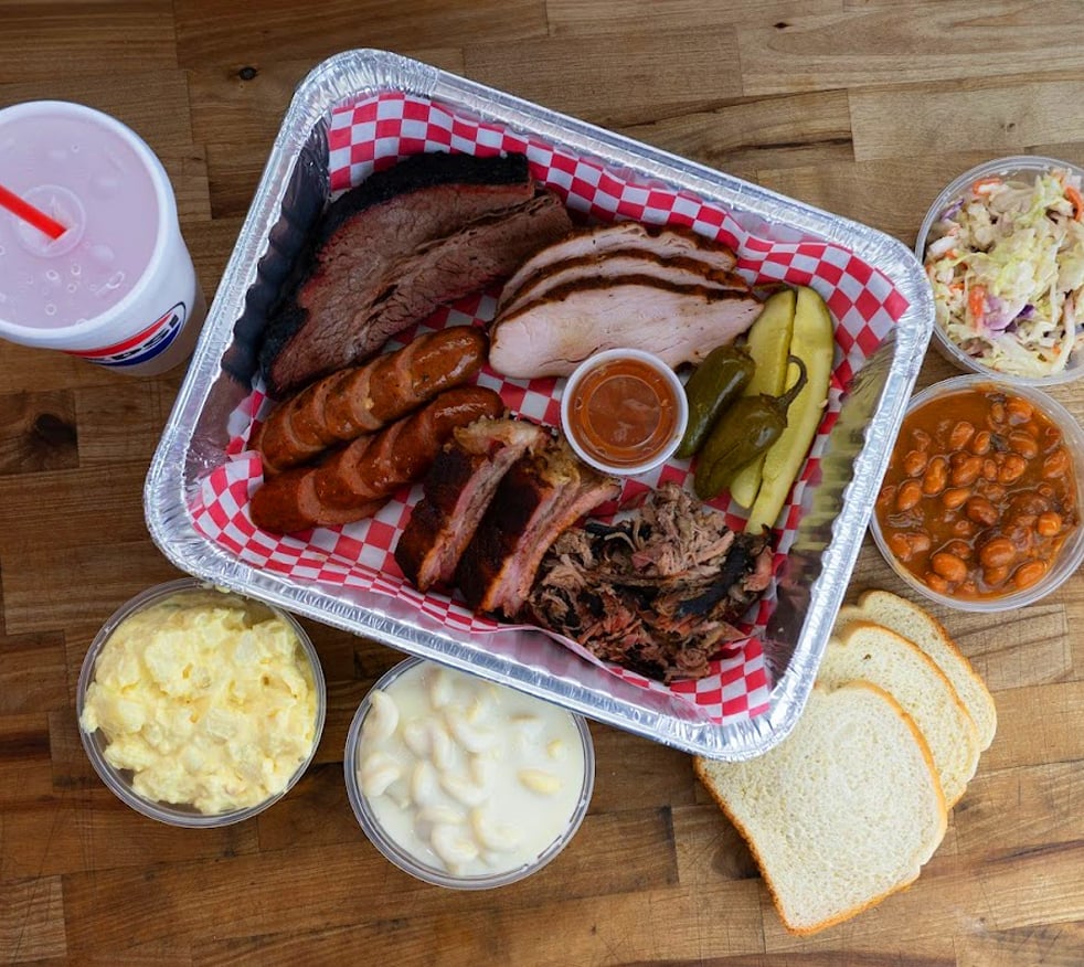 Barbecue platter with assorted smoked meats, sausages, and sides including coleslaw, potato salad, beans, and bread on a wooden table