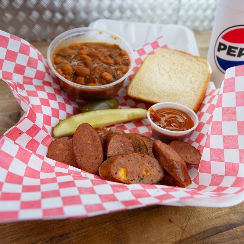 A make-it-a-meal BBQ plate with sliced sausage, beans, toast, pickle, and a Pepsi on red-checkered paper