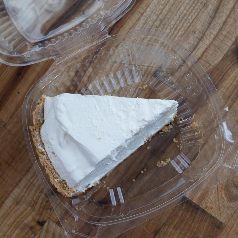 Slice of cheesecake with graham cracker crust in a clear plastic container on a wooden surface