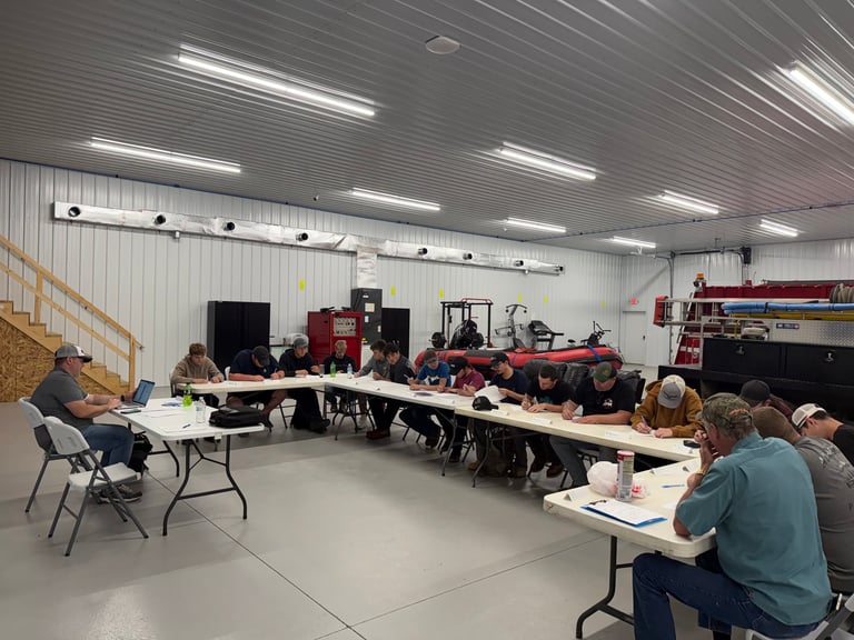Group meeting in a warehouse with people seated at long tables, farm equipment and machinery visible in the background.