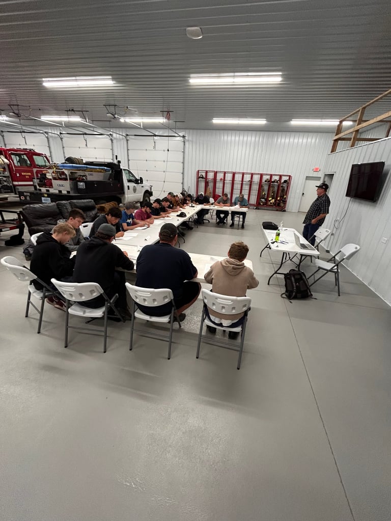 Group of people seated around tables in a large industrial warehouse space with fluorescent lighting and vehicles in background