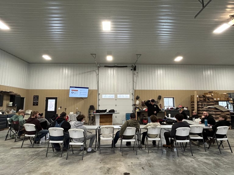 Group of people seated in white chairs arranged in rows inside a large industrial warehouse-style room with exposed ceiling and bright lighting.