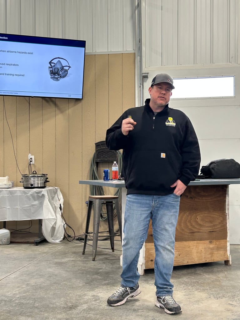 Man in black jacket and cap speaking at an indoor presentation with a display screen and equipment visible behind him