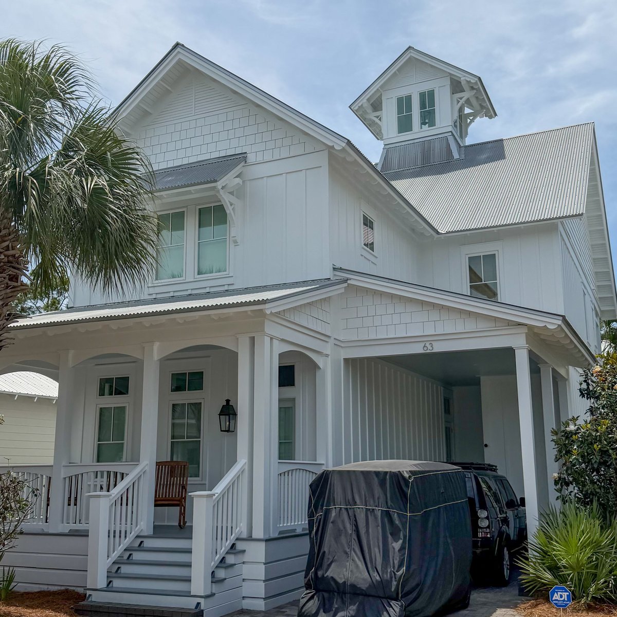 White two-story farmhouse with wrap-around porch, cupola, metal roof, and palm trees in landscaping