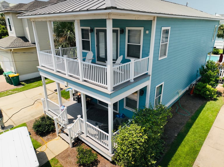 Elevated blue beach house with white railings, metal roof, two levels, and landscaped yard on clear day