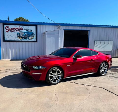 Red Mustang GT sports car parked outside Sharmies auto shop with metal building in background