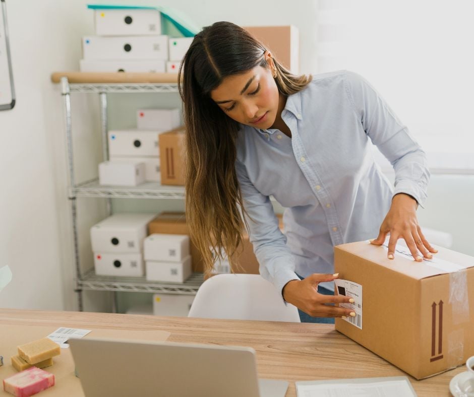 Woman packing cardboard box at desk with laptop and shelves of storage boxes in background