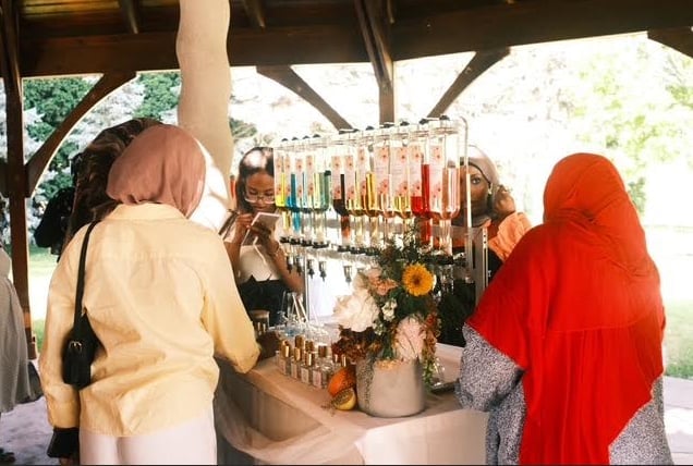 Customers browsing colorful oils and products at an outdoor market stall with wooden roof structure