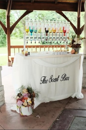 A scent bar station at an event with colorful candles displayed on shelves above a draped white table labeled The Scent Bar with flowers