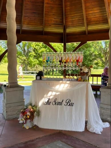 An elegant outdoor scent bar setup under a covered pavilion with hanging perfume bottles and floral arrangements