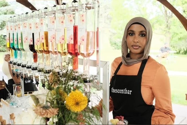 Woman wearing a black apron standing next to a display of colorful lipsticks and nail polish bottles with fresh flowers