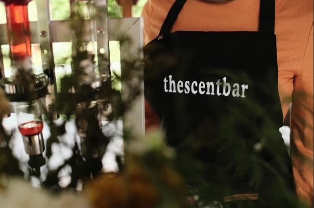 Person wearing black apron labeled theescentbar in a shop with shelves of bottled products in background