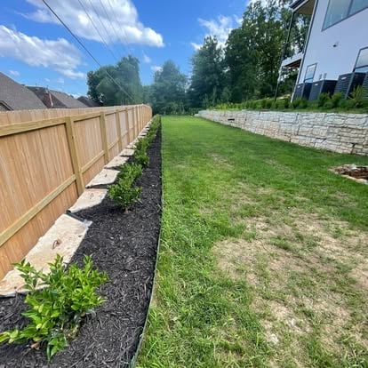 Backyard with wooden fence on left and stone wall on right, bordered by landscaping strips and lush green lawn in center
