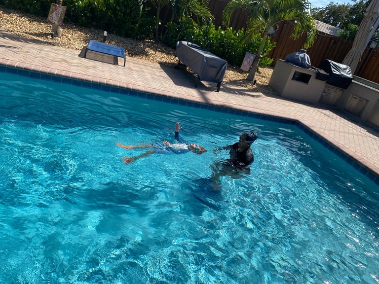 Two people swimming in a bright turquoise backyard pool on a sunny day with concrete deck and trees in background