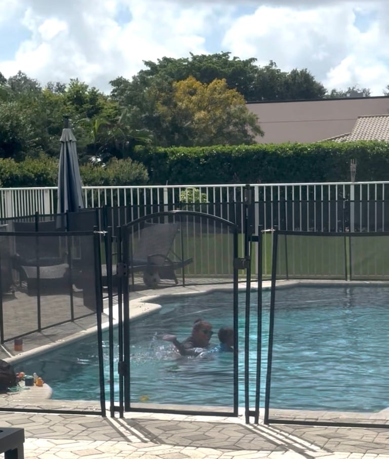 Child swimming in backyard pool with black safety gate fence, metal railings, and trees in background on sunny day