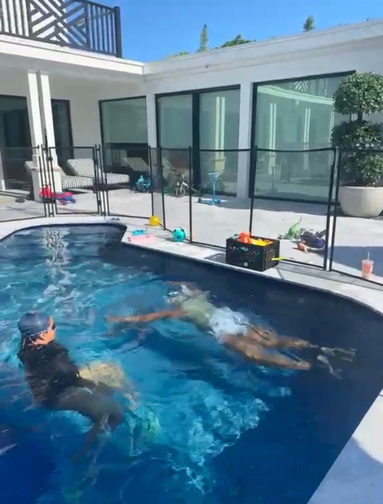 Two children swimming in a blue backyard pool with modern white house and large windows in background on a sunny day