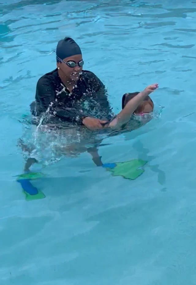 Child swimming in pool wearing black cap and goggles, using green and blue flippers