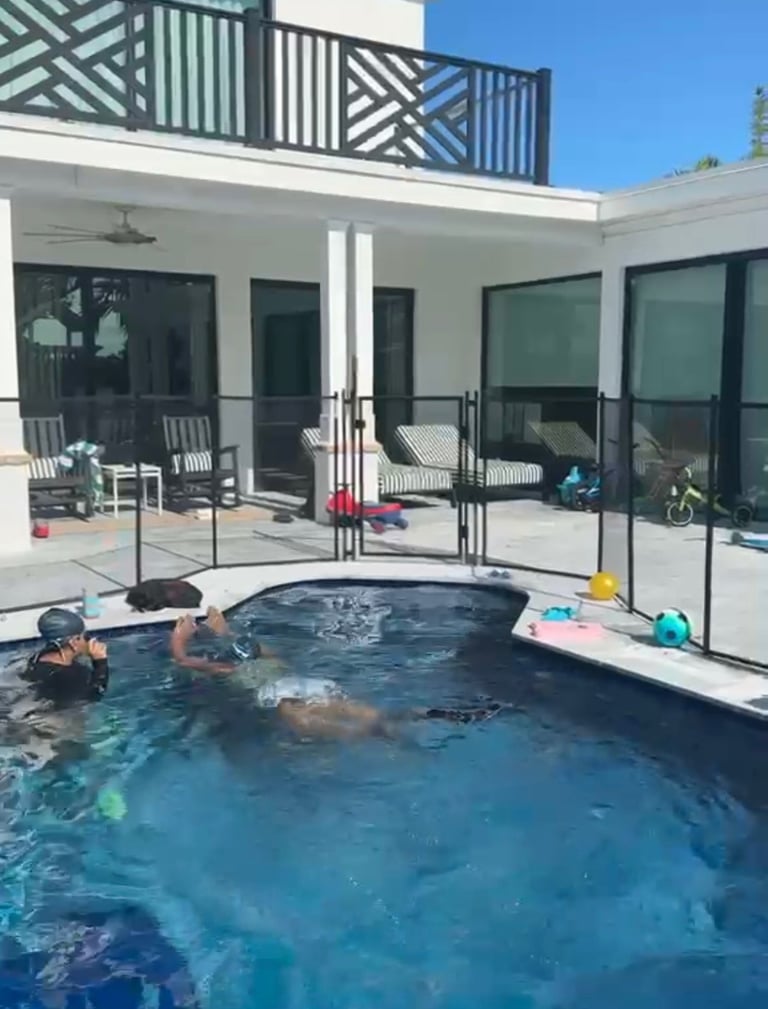 People swimming in a backyard pool next to a modern white house with large black-framed sliding glass doors and a second-story railed balcony