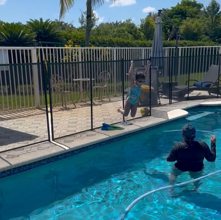 Child on pool deck reaching toward adult in swimming pool with safety fence visible and tropical landscape background