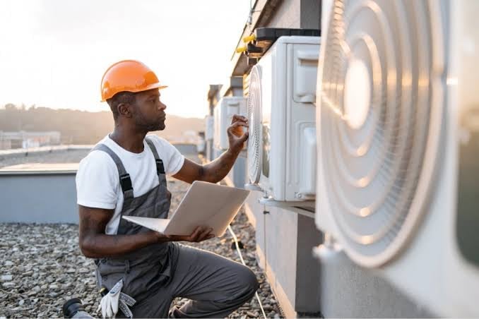 Technician in orange hard hat servicing HVAC unit on rooftop while holding tablet