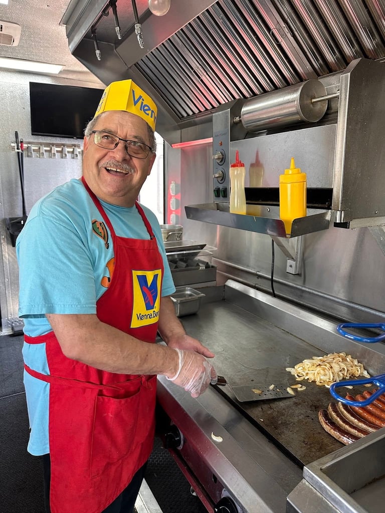 Smiling man in Vevo uniform and red apron working at food service counter with hot dog roller and condiments