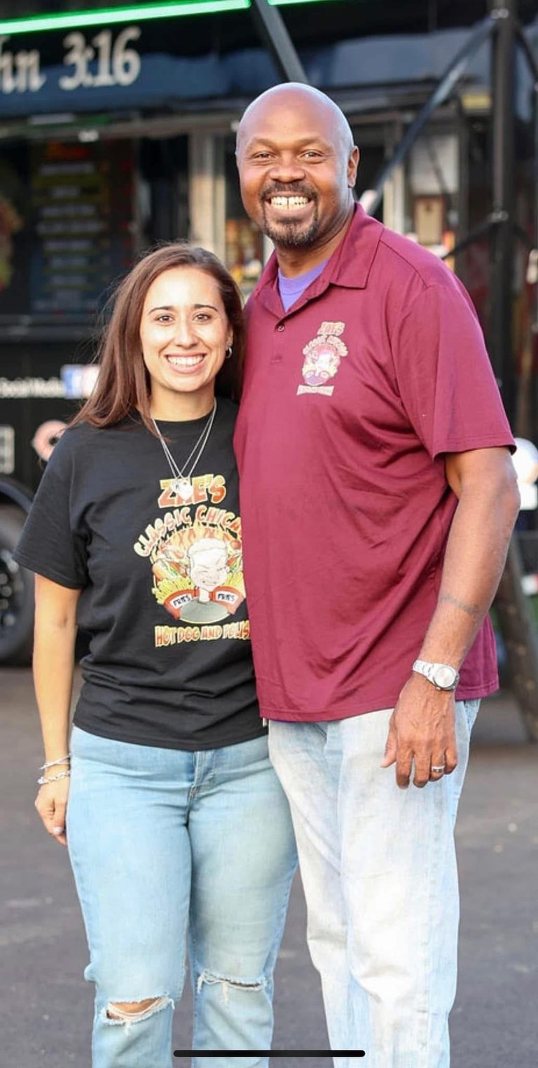 Two smiling people posing together on a city street, wearing casual clothing and smiling at the camera