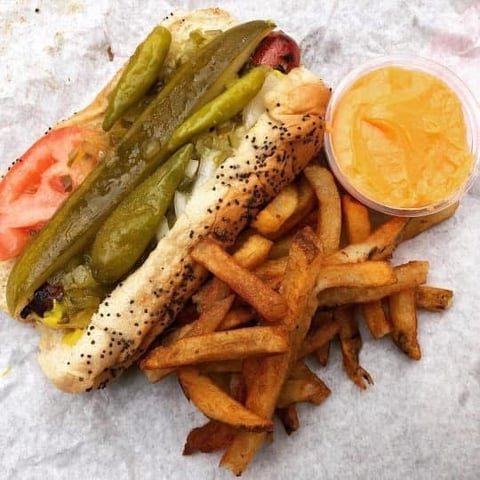 Gourmet hot dog topped with pickles and onions, served with golden fries and cheese sauce on parchment paper