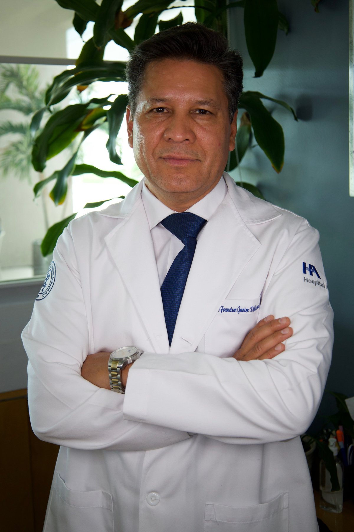 Dr. Francisco Villarreal con bata blanca y corbata azul, de pie con los brazos cruzados frente a plantas verdes