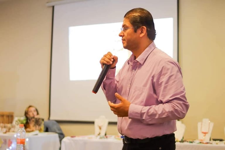 Raúl Reyes con camisa rosa hablando por micrófono frente a una audiencia con pantalla de proyección al fondo durante una presentación