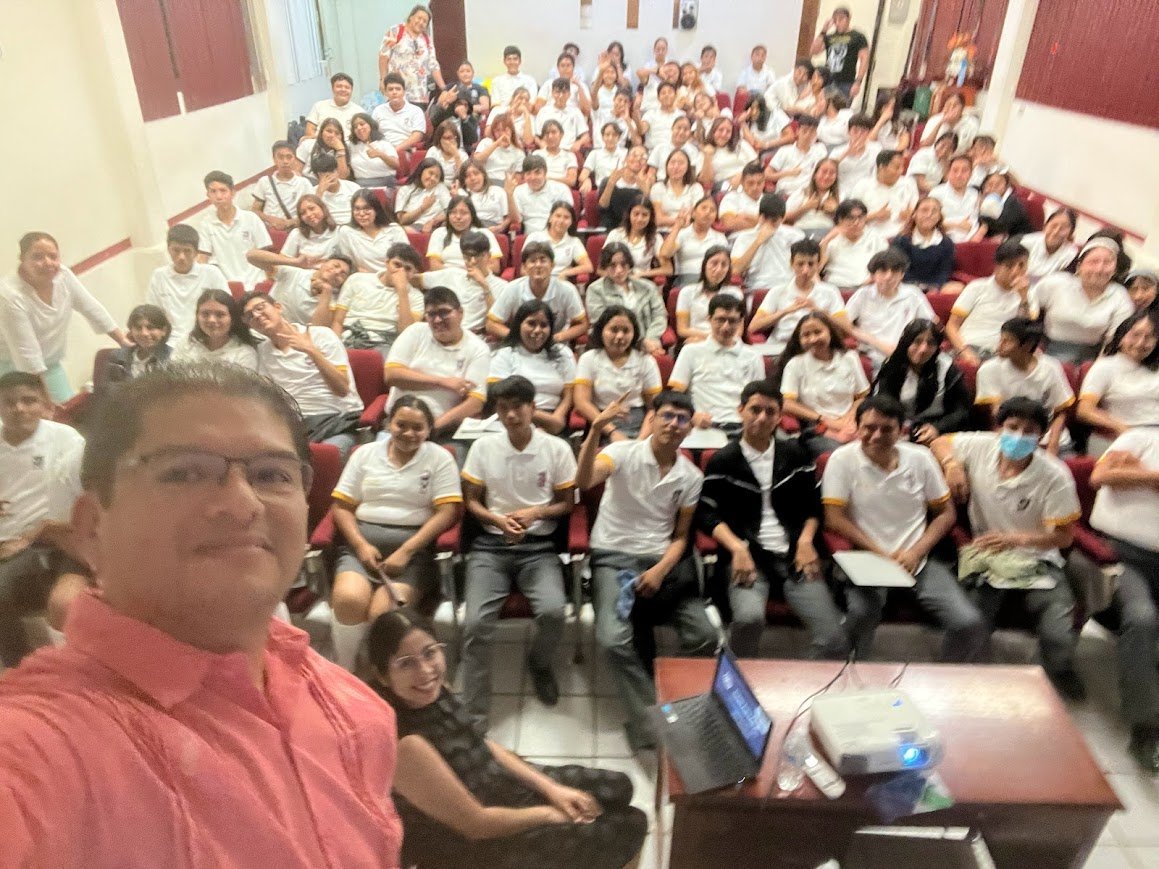 Raúl Reyes tomando selfie con un grupo grande de personas vestidas con camisas blancas en un auditorio durante una charla