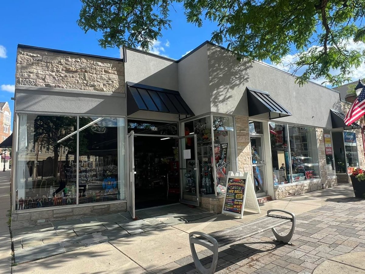 Street-facing OneStopWonders storefront with gray and stone exterior, black awnings, display windows, and a metal bench on the sidewalk
