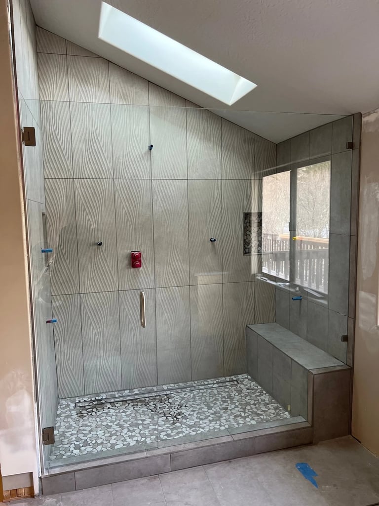 Modern walk-in shower enclosure with wavy patterned tile walls, pebble floor, glass doors, built-in bench, and skylight
