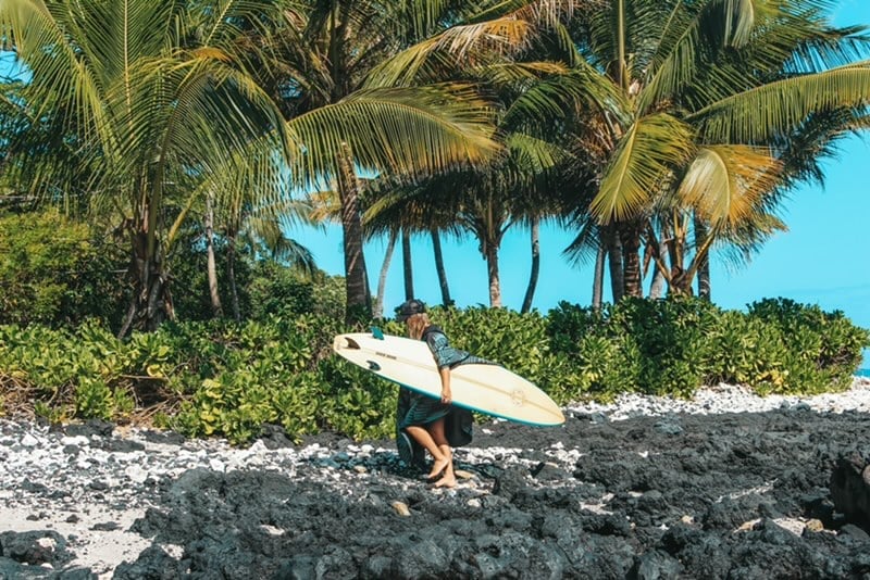 Surfer carrying white surfboard walking on rocky beach with palm trees and clear blue sky in background